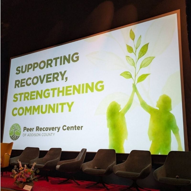 Stage at a Peer Recovery Center of Addison County event featuring a presentation screen reading “Supporting Recovery, Strengthening Community,” with green silhouettes holding a growing tree and chairs arranged for a panel discussion.